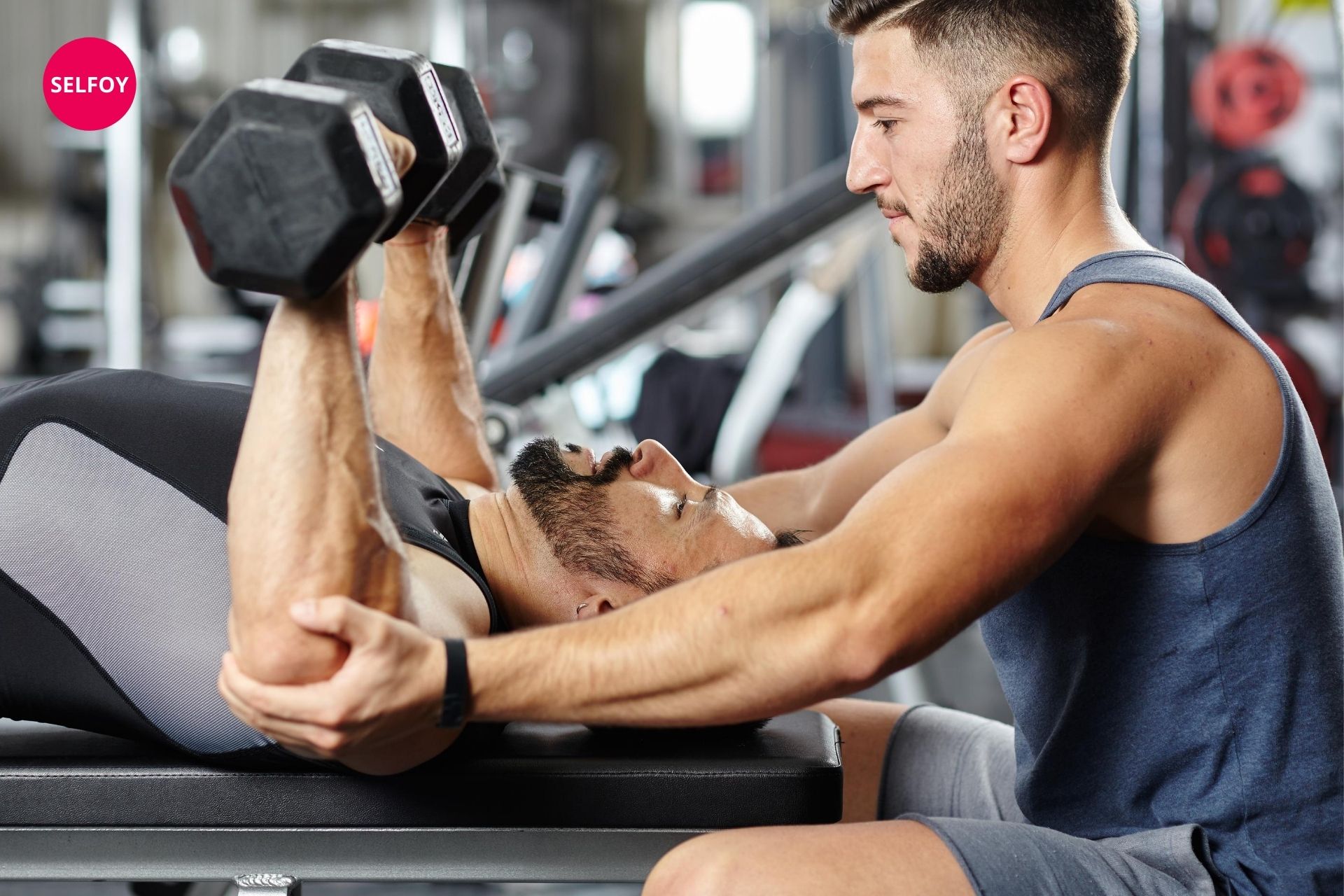 Man lifting dumbells and another guy is giving support to him at yelbow